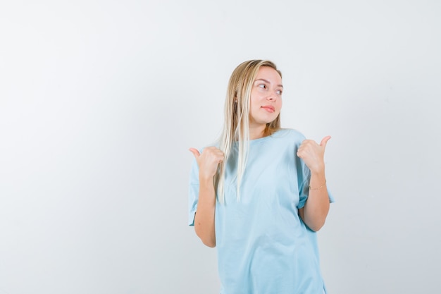 Ragazza bionda in maglietta blu che indica da parte con i pollici e che sembra felice, vista frontale.