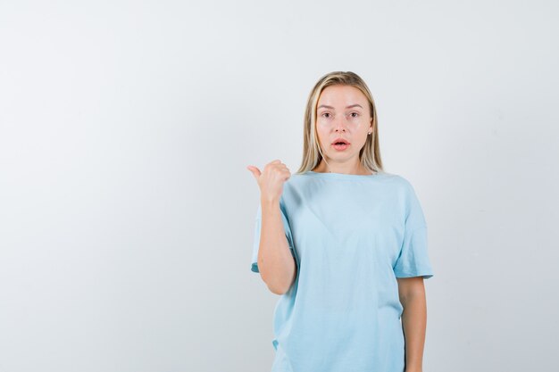 Ragazza bionda in maglietta blu che indica da parte con il pollice e che sembra sorpresa, vista frontale.