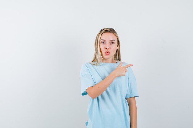 Ragazza bionda in maglietta blu che punta a destra con il dito indice e che sembra sorpresa, vista frontale.