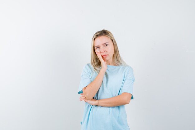 Ragazza bionda in maglietta blu che si appoggia sulla guancia sul palmo, tenendo la mano sotto il gomito e guardando piuttosto, vista frontale.