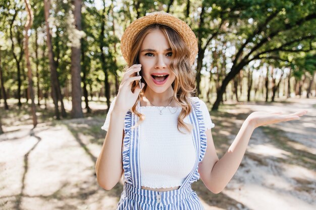 Ragazza bionda in maglietta casual parlando emotivamente al telefono. Ritratto all'aperto di donna riccia divertente in posa con lo smartphone sugli alberi.