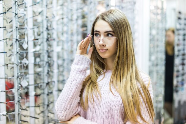 Ragazza bionda in maglione bianco sceglie nuovi occhiali medicali in negozio professionale