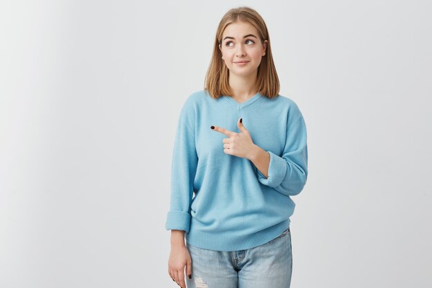 Ragazza bionda in maglione blu che guarda con i suoi occhi scuri da parte che indica con il dito indice lo spazio della copia che fa pubblicità a qualcosa. Donna che posa contro la parete grigia con lo spazio della copia per testo o promozione