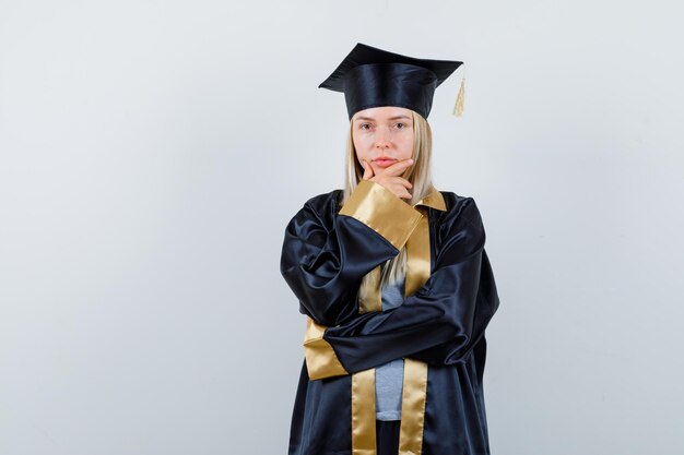 Ragazza bionda in piedi in posa di pensiero, mettendo la mano sotto il mento in abito da laurea e berretto e guardando pensierosa.