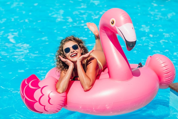 Ragazza bionda in posa per storie di Instagram, che riposa nella piscina estiva su un fenicottero rosa gonfiabile in costume da bagno.