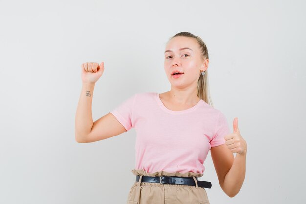 Ragazza bionda in t-shirt, pantaloni che fingono di mostrare qualcosa con il pollice in su e guardando fiducioso, vista frontale.