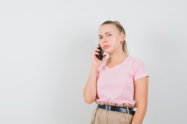 Ragazza bionda in t-shirt, pantaloni, parlando al cellulare e guardando triste, vista frontale.