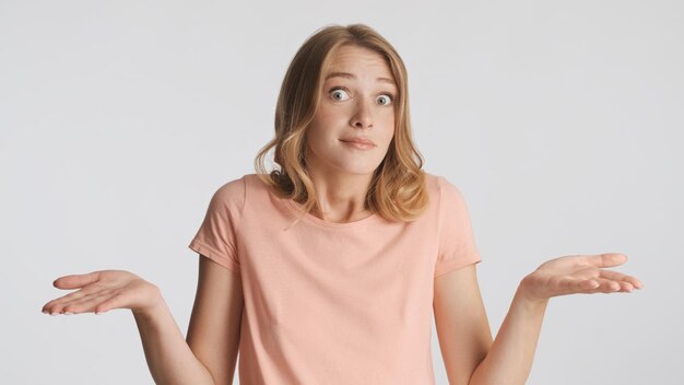 Ragazza bionda incerta che scrolla le spalle le spalle mostrando che non so gesto sulla fotocamera isolato su sfondo bianco Concetto di linguaggio del corpo