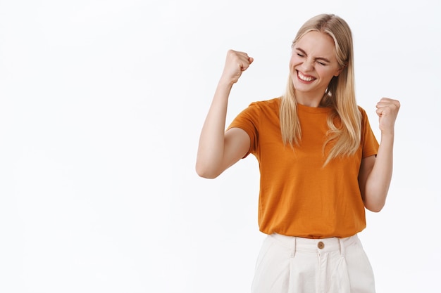 Ragazza bionda moderna di successo, elegante e incoraggiata con tatuaggi in t-shirt arancione, pompa a pugno e sorridente, dì di sì per celebrare il successo, raggiungere l'obiettivo, provare il gusto della vittoria, vincere alla lotteria