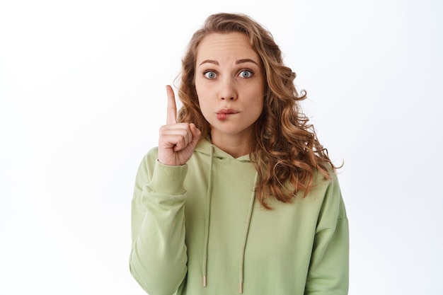 Ragazza bionda sciocca e carina che morde il labbro confuso, puntando il dito verso il logo, mostrando qualcosa di strano, in piedi contro il muro bianco