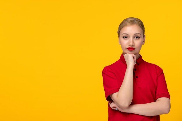 Ragazza bionda seria di giorno della camicia rossa che tiene la mano sotto il mento in una camicia rossa su sfondo giallo