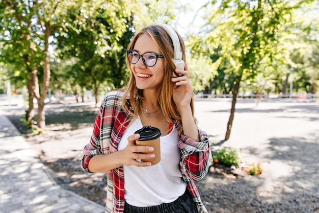 Ragazza bionda soddisfatta con gli occhiali in posa in cuffie bianche. Ritratto di adorabile giovane donna in piedi sulla natura con il tè.
