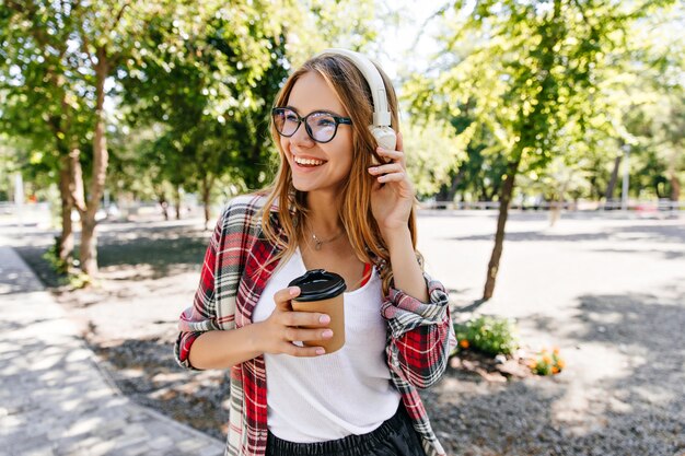 Ragazza bionda soddisfatta con gli occhiali in posa in cuffie bianche. Ritratto di adorabile giovane donna in piedi sulla natura con il tè.