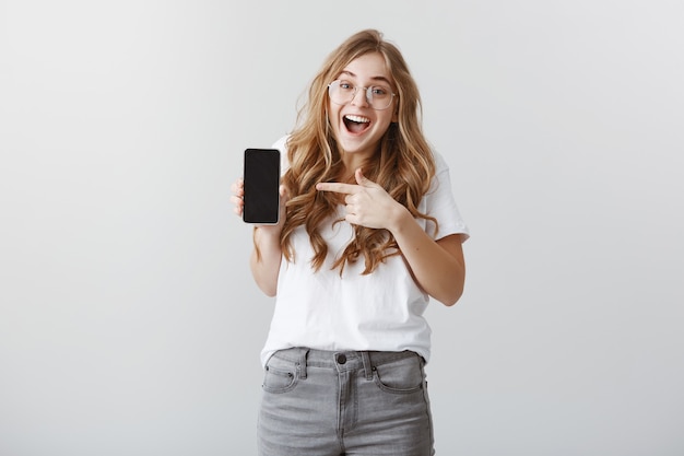 Ragazza bionda sorpresa ed eccitata con gli occhiali che punta il dito sul display del telefono cellulare, mostrando l'applicazione