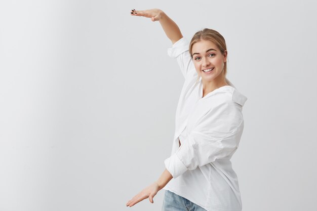 Ragazza bionda stupita in camicia bianca che mostra con la lunghezza delle mani della scatola. Abbastanza femminile che indossa abiti casual sorridente con i denti, dimostrando la dimensione di qualcosa di grande.