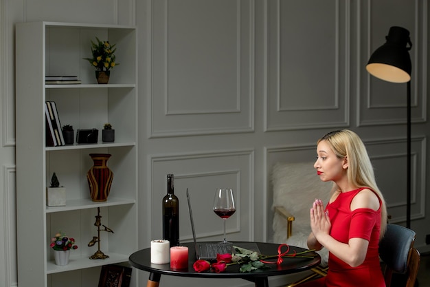 Ragazza bionda sveglia di amore virtuale in vestito rosso alla data di distanza con il vino che prega le mani insieme