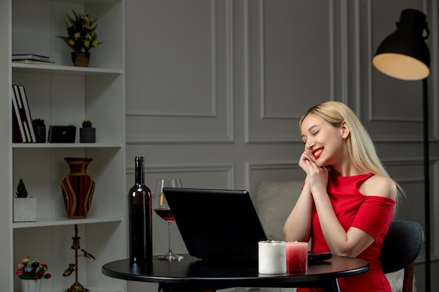 Ragazza bionda sveglia di amore virtuale in vestito rosso alla data di distanza con il vino felice