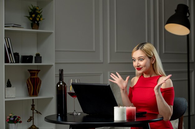 Ragazza bionda sveglia di amore virtuale in vestito rosso alla data di distanza con il vino sorpreso