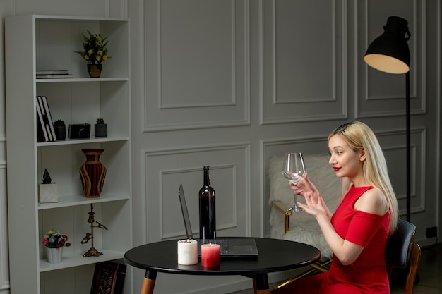 Ragazza bionda sveglia di amore virtuale in vestito rosso alla data di distanza con vino e candele che tengono il vetro