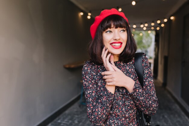 Ragazza bruna animata in berretto rosso e abito vintage si strofina le mani, decidendo di prendere in giro un amico. Ritratto del primo piano di giovane donna eccitata con capelli corti scuri e sorriso di hollywood che guarda lontano