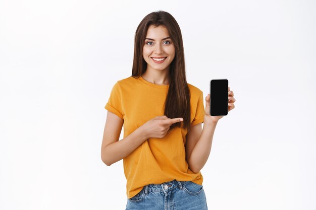 Ragazza bruna attraente allegra e sicura di sé in maglietta gialla, con in mano lo smartphone, puntando lo schermo mobile e sorridendo, consiglia un'applicazione telefonica fantastica, dà il collegamento al codice promozionale, omaggio