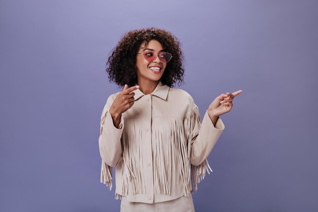 Ragazza bruna con occhiali lilla e abito beige mostra il dito a lato Donna felice in giacca elegante sorridente su sfondo viola