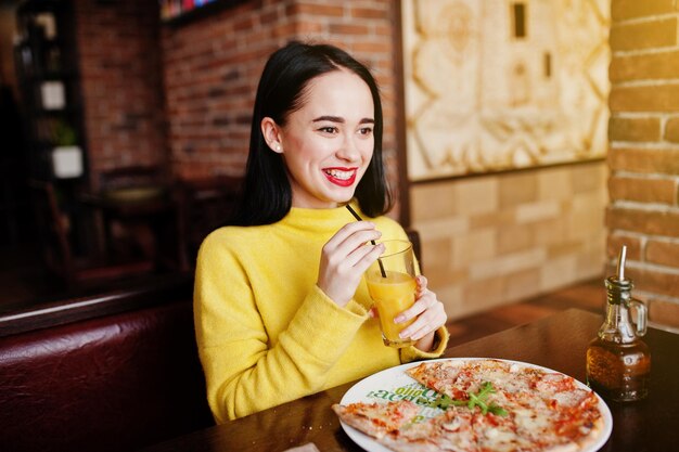 Ragazza bruna divertente in maglione giallo che mangia pizza al ristorante