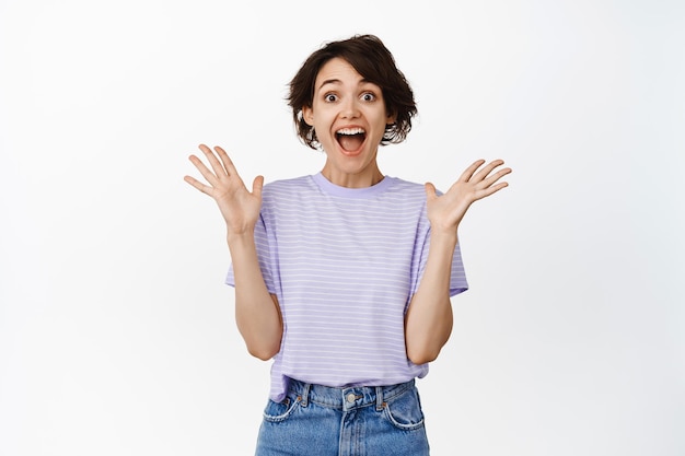 Ragazza bruna eccitata e sorpresa che vince, urlo felice, batti le mani e guarda in soggezione, celebrando la vittoria, trionfando di gioia, in piedi su sfondo bianco.