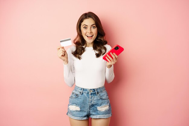 Ragazza bruna entusiasta che mostra la carta di credito e utilizza il telefono cellulare per ordinare o pagare, app per lo shopping online, in piedi su sfondo rosa