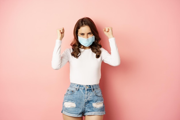 Ragazza bruna entusiasta in maschera medica, ballando e ridendo, celebrando la vittoria, trionfando, vincendo smth, in piedi su sfondo rosa