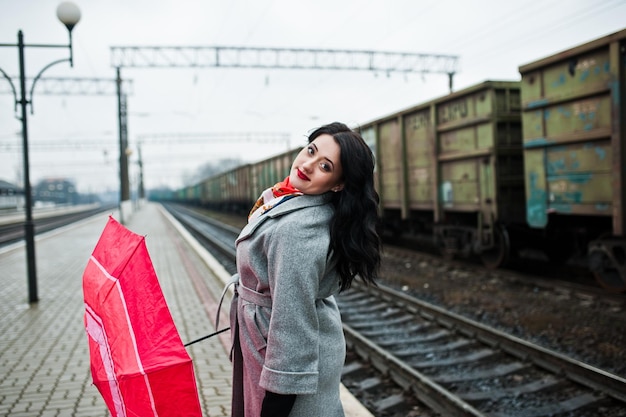 Ragazza bruna in cappotto grigio con ombrello rosso nella stazione ferroviaria