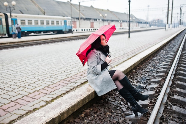 Ragazza bruna in cappotto grigio con ombrello rosso nella stazione ferroviaria