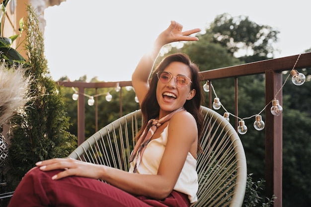 Ragazza bruna in occhiali rosa pone sul balcone e mostra il segno di pace. Donna allegra in sciarpa di seta intorno al collo ride e si siede sulla terrazza.