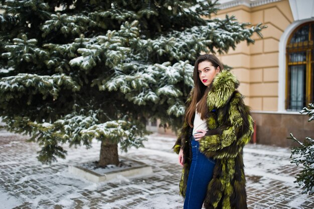 Ragazza bruna in pelliccia verde al giorno d'inverno contro il pino innevato