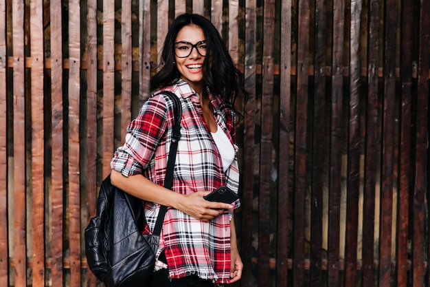 Ragazza bruna ispirata con il telefono in posa sulla parete di legno con un sorriso allegro. Ritratto all'aperto di affascinante signora latina in occhiali alla moda.