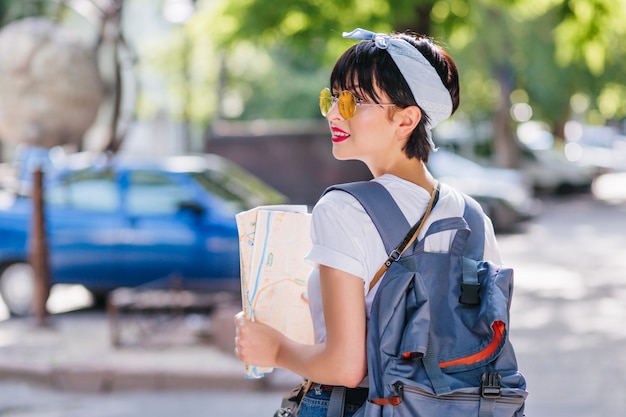 Ragazza bruna leggermente abbronzata indossa accessori alla moda guardandosi intorno perplessa, tenendo in mano la mappa della città