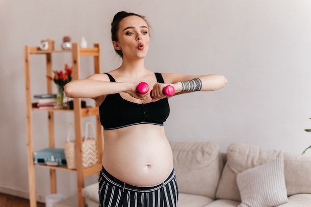 Ragazza bruna respira profondamente e fa esercizi sportivi. La donna incinta nella parte superiore nera tiene i manubri rosa nel soggiorno.