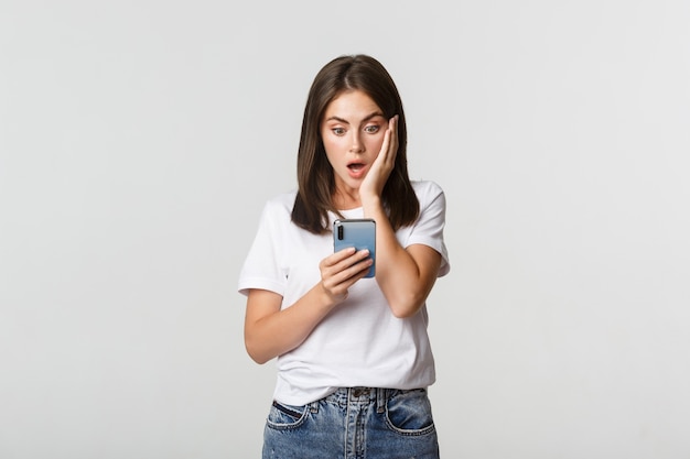 Ragazza bruna scioccata e colpita guardando stupito lo schermo dello smartphone.