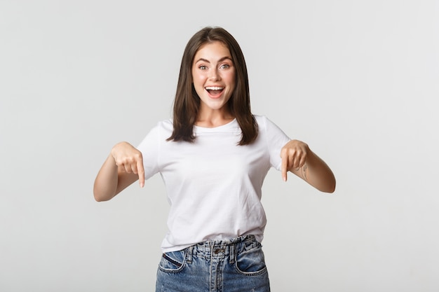 Ragazza bruna sorridente soddisfatta che punta le dita verso il basso, consiglia il prodotto.