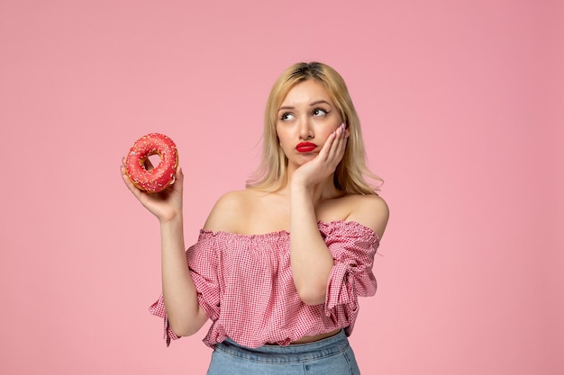 Ragazza carina adorabile pulcino con rossetto rosso in camicetta rosa pensando a qualcosa che tiene la ciambella