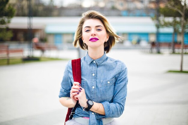 Ragazza carina che cammina con capelli biondi corti e volanti e labbra rosa brillante che tiene il suo zaino di marsala sulla spalla e indossa una camicia di jeans blu e una gonna di tulle grigia.