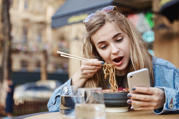 Ragazza carina che mangia spaghetti cinesi e guardando il telefono, tenendo c