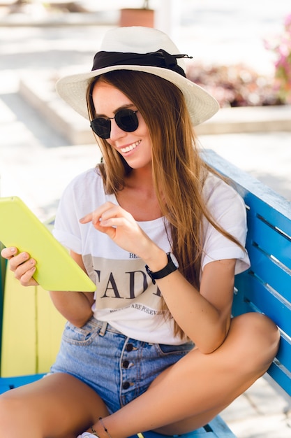 Ragazza carina che si siede su una panchina blu e gioca su un tablet in custodia gialla.