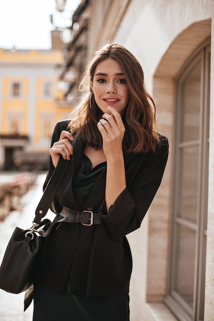 Ragazza carina con acconciatura ondulata media, trucco moderno, abito di seta verde, giacca nera e accessori eleganti in posa sulla strada diurna e guardando davanti