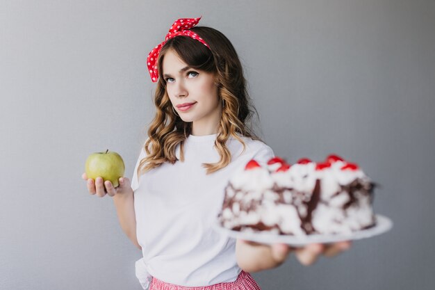 Ragazza carina con capelli mossi scuri pensando a calorie e tenendo la torta. Foto interna di bella giovane donna con torta di mele e cremosa al cioccolato.