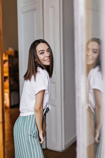 Ragazza carina con capelli scuri rilassante a casa e sorridente. Foto dell'interno della donna europea positiva in pigiama che ride mattina.