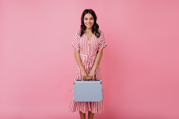 Ragazza carina con lunghi capelli scuri in abito rosa modestamente in posa con valigetta blu, guardando con un sorriso amichevole.