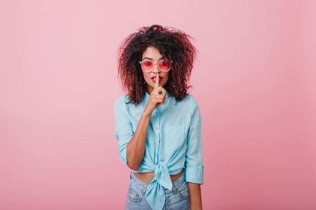 Ragazza carina e magra richiede silenzio. Ritratto dell'interno della signora africana alla moda che dice un segreto.