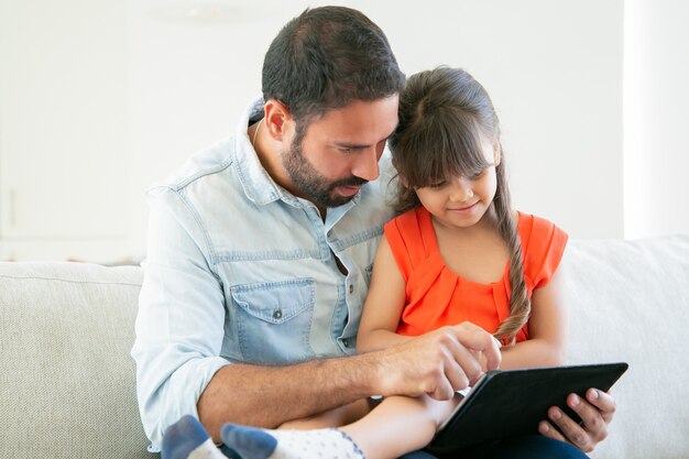 Ragazza carina e suo padre che guardano film o leggono insieme sullo schermo del tablet.