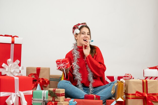 Ragazza carina festa di vista frontale con il regalo della holding del cappello della Santa usando il noisemaker che si siede intorno ai regali